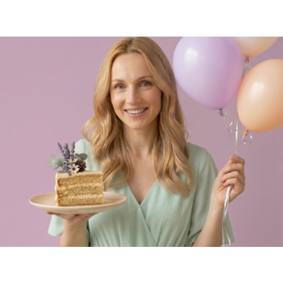 Uśmiechnięta kobieta o jasnych włosach trzymająca w dłoni kawałek tortu i balony - odkryj najlepsze naturalne kosmetyki tołpa.®z okazji naszych urodzin.