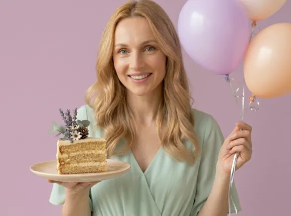 Uśmiechnięta kobieta o jasnych włosach trzymająca w dłoni kawałek tortu i balony - odkryj najlepsze naturalne kosmetyki tołpa.®z okazji naszych urodzin.