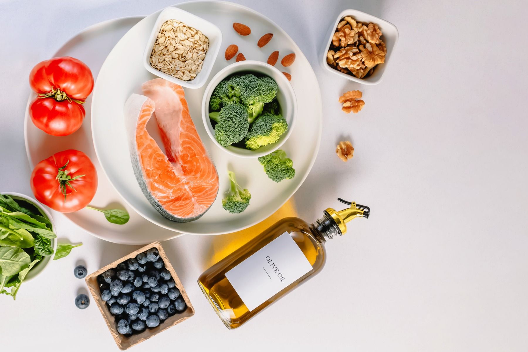 Na białym tle rozłożone różne zdrowe i pożywne produkty spożywcze takie jak oliwa z oliwek, orzechy włoskie, pomidory, płatki owsiane, brokuły, łosoś, borówka amerykańska czy szpinak